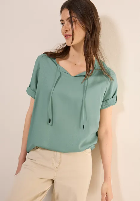 Frau mit mintgrüner Bluse und heller Hose steht an einer Wand.